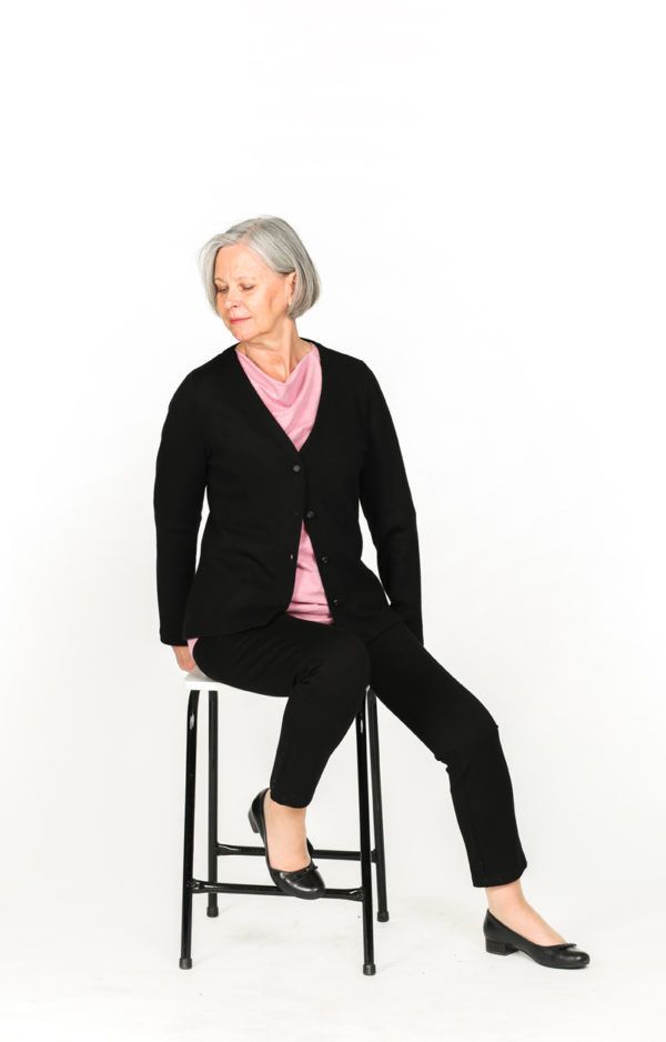 Kuvassa malli istuu jakkaralla päällään musta SOFIA-merinovillaneuletakki. Neuletakki on yhdistetty mustiin housuihin ja vaaleanpunaiseen paitaan. Neuletakki on ylhäältä kiinni hakaskiinnityksellä.