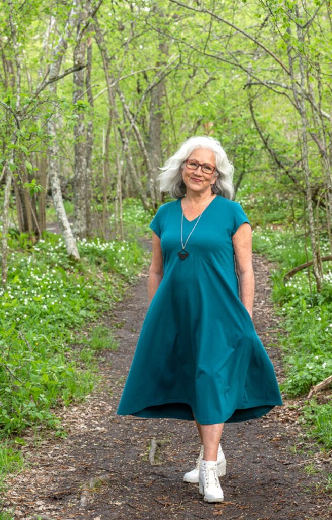 Kuvassa on vehreän metsätaustan edessä iloinen malli, jolla on päällään SUVI-mekko pikkuhihoilla smaragdin vihreänä. SUVI-mekko on V-aukkoinen ja siinä on leveä, hulmuava helma ja yläosassa imartelava leikkaus rintojen alla.